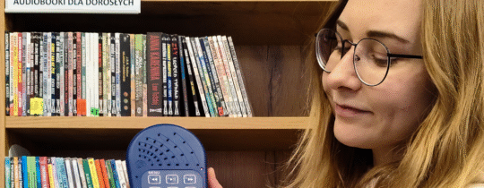 Bibliotekarka w okularach stoi na tle audiobooków i prezentuje w rękach Czytak.